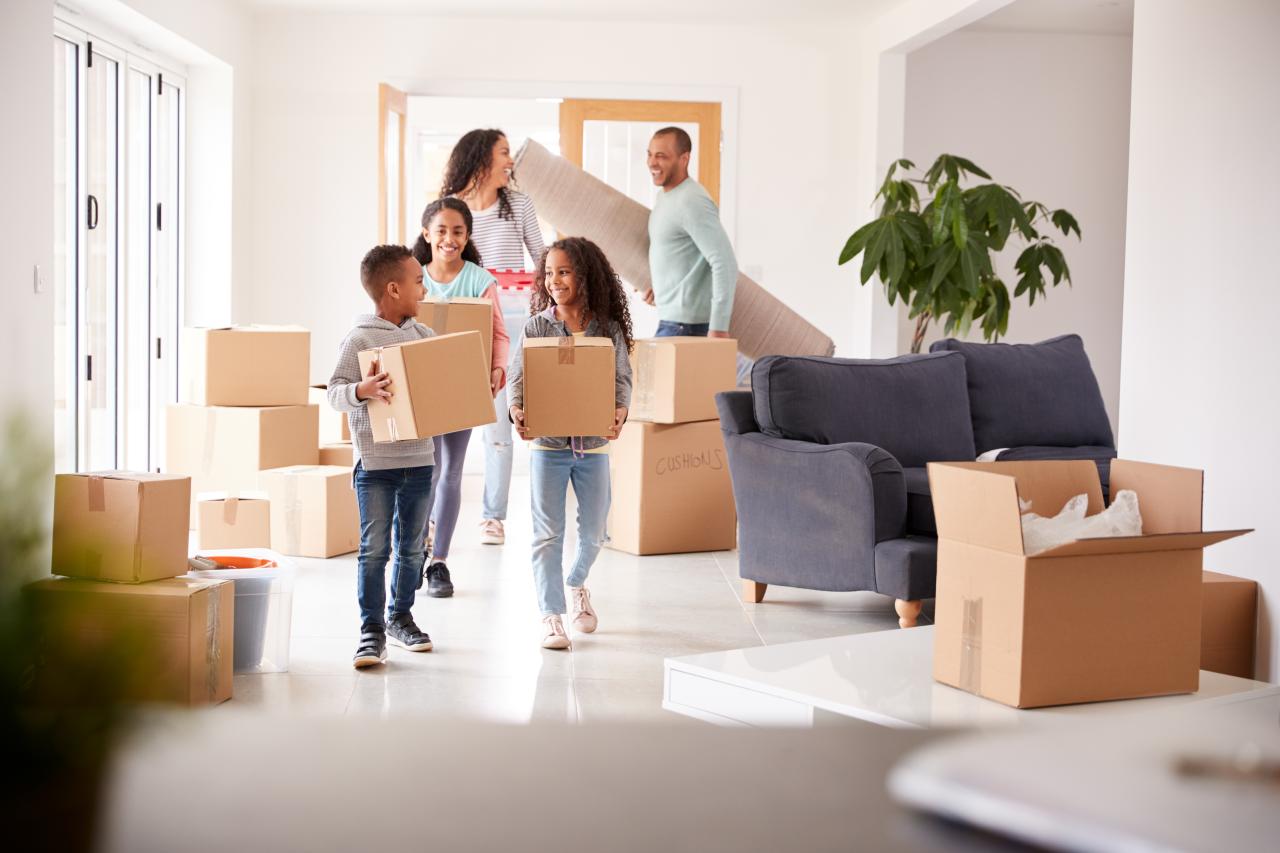 An image of a family of four carrying boxes and a rug into a new, home. They are all smiling.